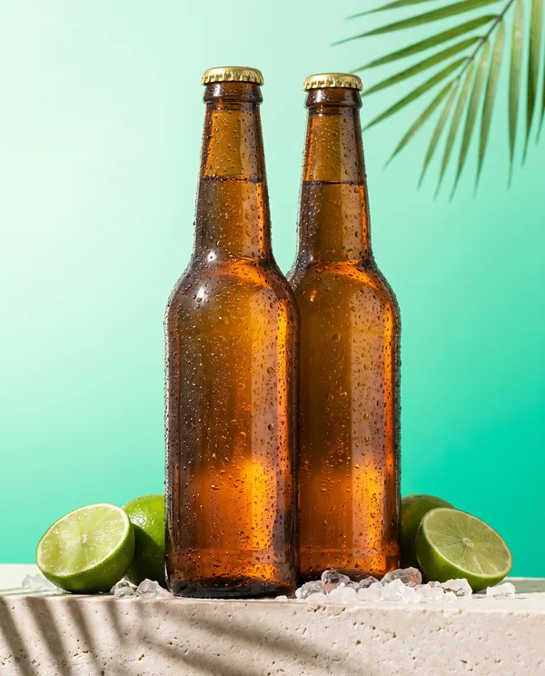 Dos botellas de cerveza ámbar con condensación sobre hielo, con medias limas, en fondo verde menta