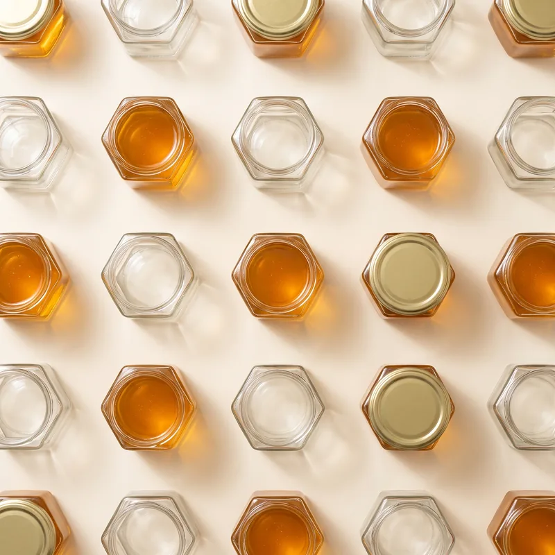 Retícula de frascos hexagonales vistos desde arriba sobre fondo crema, unos llenos de miel y otros vacíos, con tapas doradas