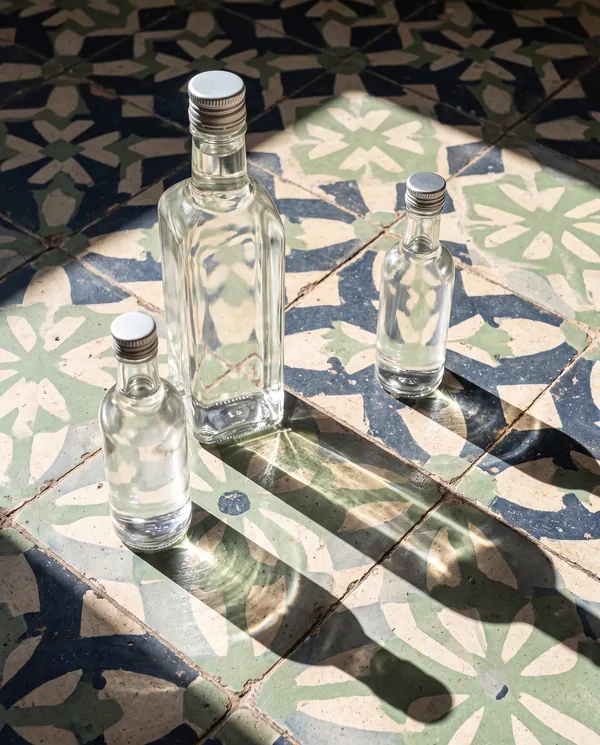Tres botellas de vidrio sobre un piso de pasta de cemento yucateca en azul marino, verde oliva y crema, con sombras largas de sol de tarde