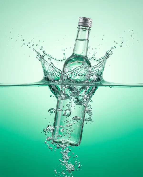 Botella de vidrio entrando en el agua y levantando una corona de salpicadura, sobre fondo verde esmeralda